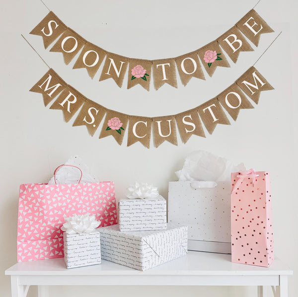 A two tiered burlap banner. The first tier reads soon to be with pink rose flowers between the words. The second tier reads mrs. custom with a pink rose flower between the words.