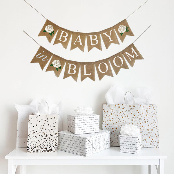 A Baby in Bloom baby shower banner in white letters on burlap flags. Starting with a white hand stenciled rose, the first line says baby with a white flower after the word, the second line reads in bloom with a white rose in between the words.