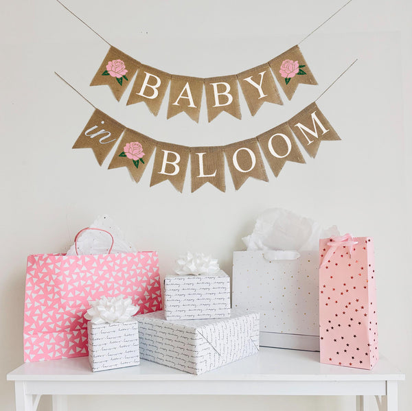 A Baby in Bloom baby shower banner in white letters on burlap flags. Starting with a pink hand stenciled rose, the first line says baby with a pink flower after the word, the second line reads in bloom with a pink rose in between the words.