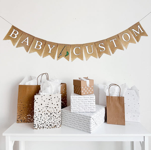 A burlap banner made from 4.5 by 6.5 inch fishtail shaped flags. Each flag has a pocket across the top for stringing. This banner says BABY CUSTOM in white capital letters. There is a hand stenciled image of a fishing rod and a green fish between the words. The banner can be personalized with a custom word or name of your choice. It is strung on a jute string that is 110 inches in length.