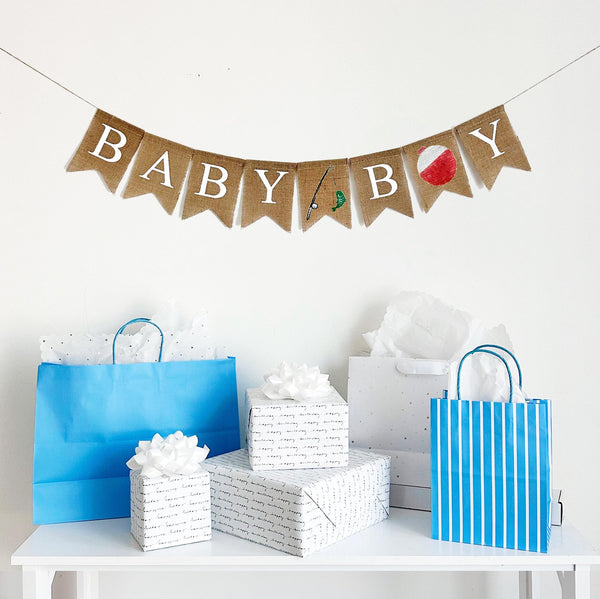 A burlap banner made from 4.5 by 6.5 inch fishtail shaped flags. Each flag has a pocket across the top for stringing. This banner says BABY BOY in white capital letters. There is a hand stenciled image of a fishing rod and a green fish between the words. Instead of the letter O, there is a hand stenciled image of a red and white bobber. It is strung on a jute string that is 110 inches in length.