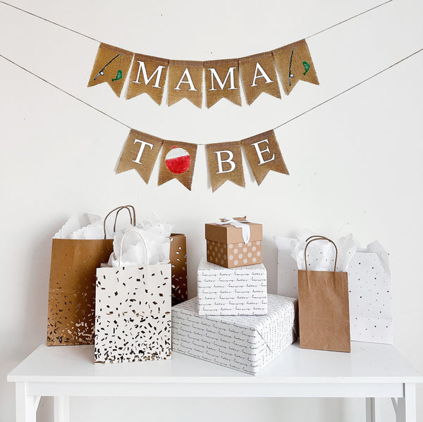 A burlap banner made from 4.5 by 6.5 inch fishtail shaped flags. Each flag has a pocket across the top for stringing. The top line says MAMA in white capital letters. There is a hand stenciled image of a fishing rod and a green fish on each end of the word. The bottom line says TO BE in white capital letters. Instead of the letter O, there is a hand stenciled image of a red and white bobber. It is strung on two jute strings that are 110 inches in length.