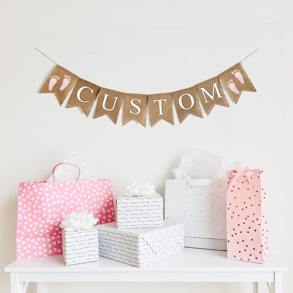 A burlap banner with the wording CUSTOM between two pink footprint image flags.