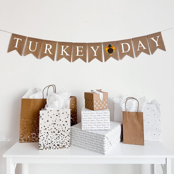 A burlap banner made from 4.5 by 6.5 inch fishtail shaped flags. The banner says TURKEY DAY in white capital letters. There is a hand stenciled image between the words of a brown turkey with orange and yellow feathers wearing a black hat. It comes with a jute string for stringing.