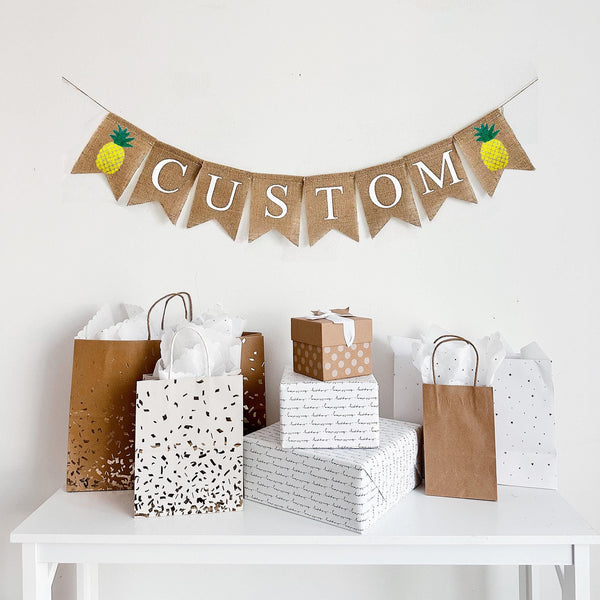 A burlap banner with hand painted pineapples at the beginning and end of the word CUSTOM.  The pineapples are painted in yellow with a green stem.