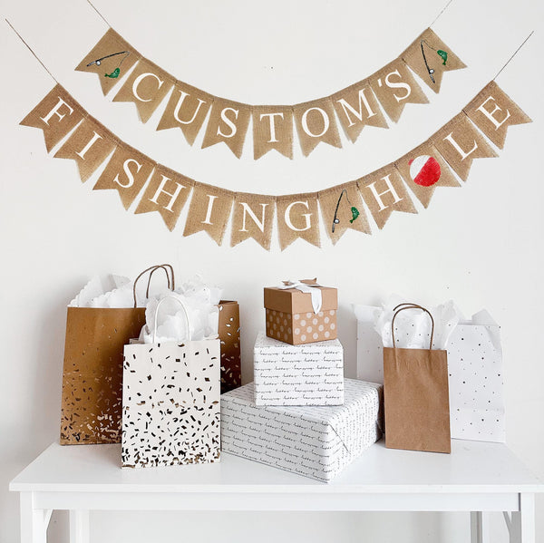 A burlap banner made from 4.5 by 6.5 inch fishtail shaped flags. Each flag has a pocket across the top for stringing. The top line is in white capital letters and can be customized with a name. There is a hand stenciled image of a fishing rod and green fish on each end of the word. The bottom line says FISHING HOLE in white capital letters. There is a hand stenciled image of a fishing rod and a green fish between the words and there is a red and white bobber instead of the letter O.