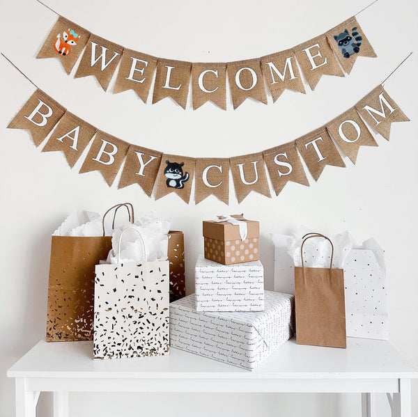 A burlap banner made from 4.5 by 6.5 inch fishtail shaped flags. Each flag has a pocket across the top for stringing. The top line says WELCOME in white capital letters. There is a hand stenciled image of an orange fox with a bow on one end and a gray raccoon on the other end. The bottom string says BABY CUSTOM and has an image of a brown squirrel between the words. Add your own name to this banner. It is strung on two jute strings that are 110 inches in length.