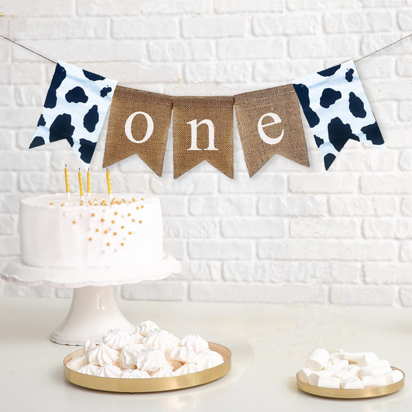 This farm themed first birthday burlap banner is make up of 5 burlap flags. The first flag is a flag made of cow print fabric followed by three more flags spelling out the word one in lowercase white letters. The banner ends with another a flag made of cow print fabric.