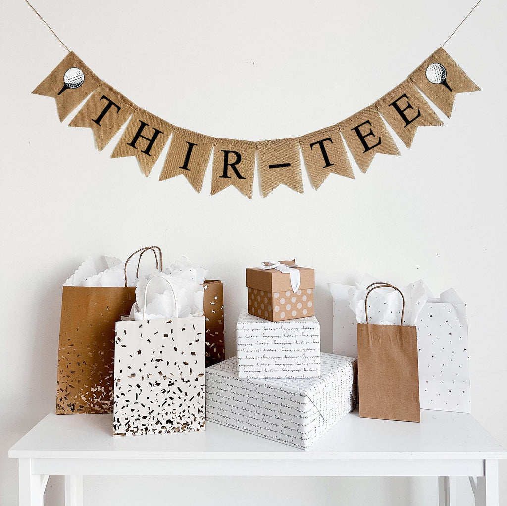 A burlap banner made from 4.5 by 6.5 inch fishtail shaped flags. Each flag has a pocket across the top for stringing. It says THIR-TEE in black capital letters and has a hand stenciled image of a white golf ball sitting on a black tee on each end. It is strung on a jute string that is 110 inches in length.
