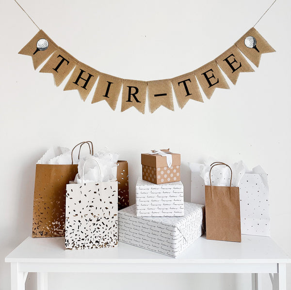 A burlap banner made from 4.5 by 6.5 inch fishtail shaped flags. Each flag has a pocket across the top for stringing. It says THIR-TEE in black capital letters and has a hand stenciled image of a white golf ball sitting on a black tee on each end. It is strung on a jute string that is 110 inches in length.