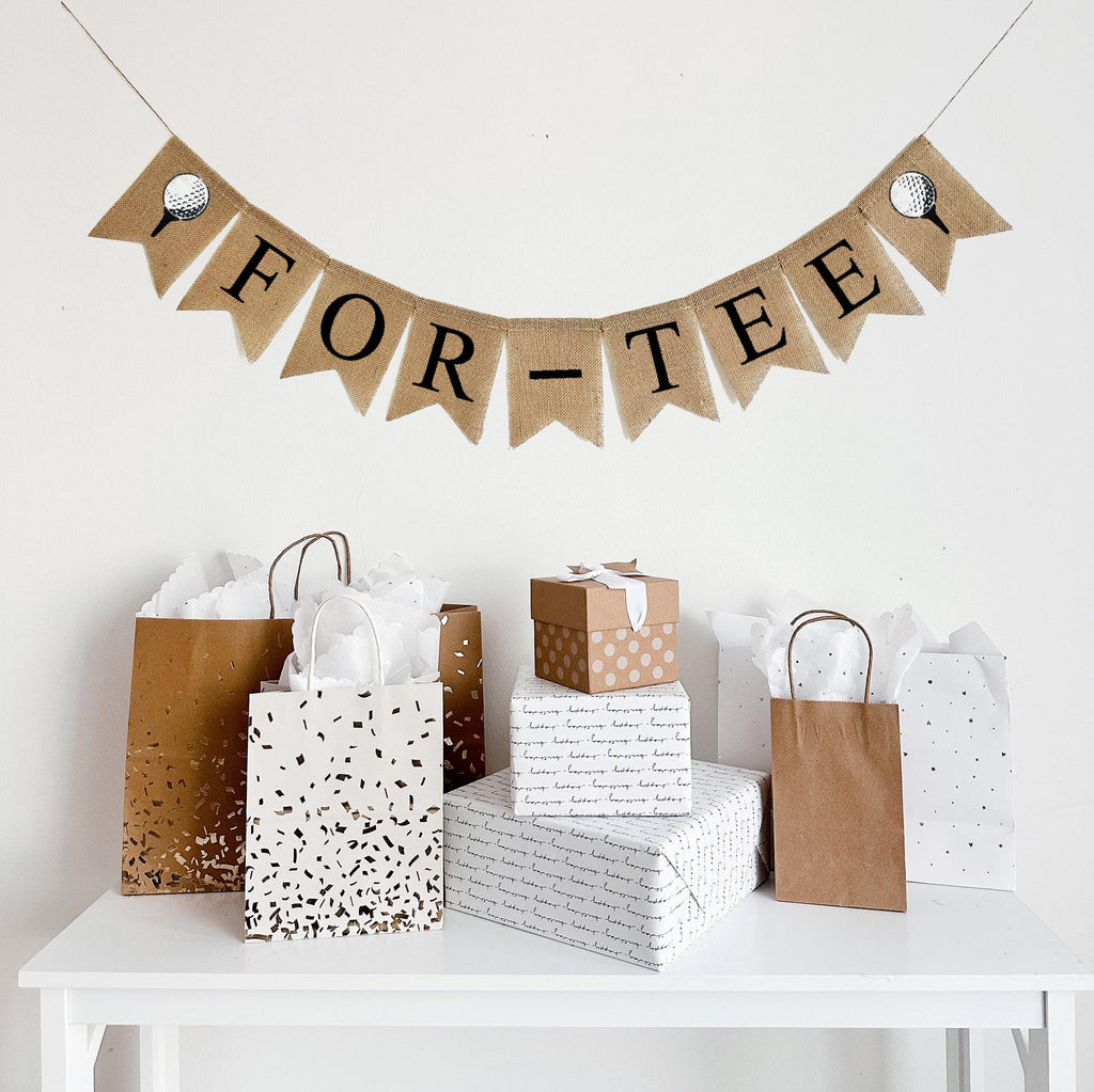 A burlap banner made from 4.5 by 6.5 inch fishtail shaped flags. Each flag has a pocket across the top for stringing. It says FOR-TEE in black capital letters and has a hand stenciled image of a white golf ball sitting on a black tee on each end. It is strung on a jute string that is 110 inches in length.