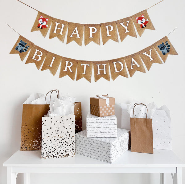 A burlap banner made from 4.5 by 6.5 inch fishtail shaped flags. Each flag has a pocket across the top for stringing. This is a custom banner in white capital letters. The top string says HAPPY and has a hand stenciled image of a fox on each end. The bottom string says BIRTHDAY and has a hand stenciled image of a raccoon on each end. It is strung on a jute string that is 110 inches in length.