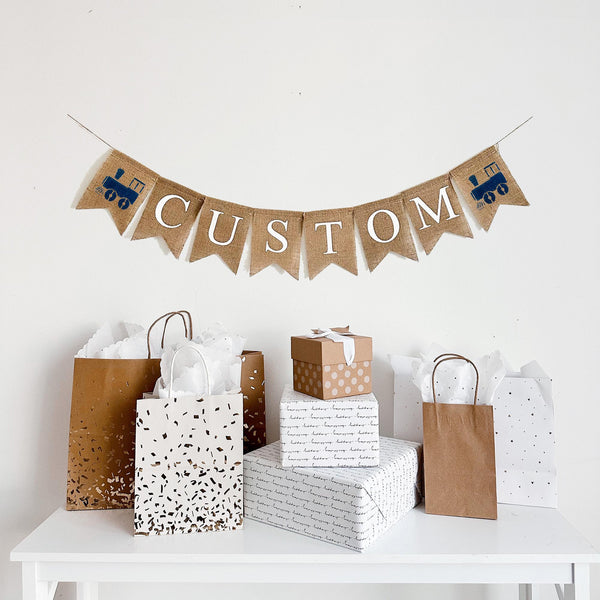 A burlap banner made from 4.5 by 6.5 inch fishtail shaped flags. Each flag has a pocket across the top for stringing. This is a custom banner in white capital letters. Add the name of your choosing to personalize this banner. There is a hand stenciled image of a navy blue train on each end of the name. It is strung on a jute string that is 110 inches in length.