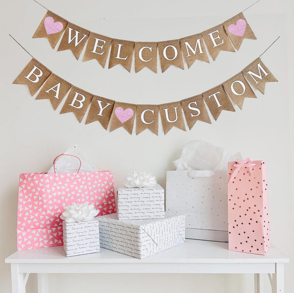 A burlap banner made from 4.5 by 6.5 inch fishtail shaped flags. Each flag has a pocket across the top for stringing. The top line says WELCOME in white capital letters. There is a hand stenciled image of a baby pink heart on each end. The bottom string says BABY CUSTOM and has a hand stenciled image of a baby pink heart between the words. Add your own name to this banner. It is strung on two jute strings that are 110 inches in length.