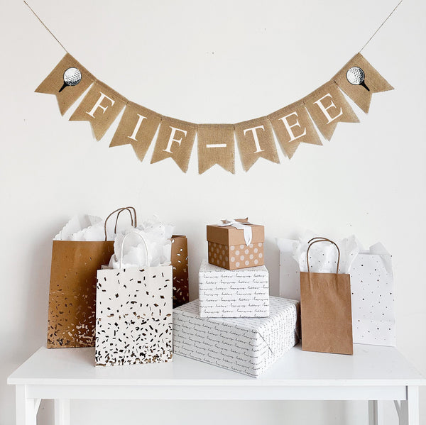 A burlap banner made from 4.5 by 6.5 inch fishtail shaped flags. Each flag has a pocket across the top for stringing. It says FIF-TEE in white capital letters and has a hand stenciled image of a white golf ball sitting on a black tee on each end. It is strung on a jute string that is 110 inches in length.