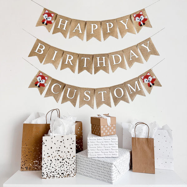 A burlap banner made from 4.5 by 6.5 inch fishtail shaped flags. Each flag has a pocket across the top for stringing. This is a custom banner in white capital letters. The top string says HAPPY and has a hand stenciled image of a fox on each end. The second string says BIRTHDAY and has no images. The bottom string can be customized with a name and has a hand stenciled image of a fox on each end. It is strung on a jute string that is 110 inches in length.