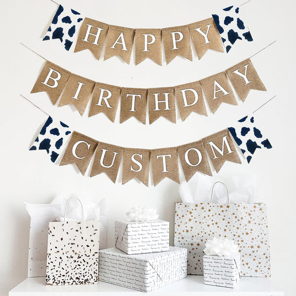 Happy Birthday banner made of cow print fabric and burlap. The first line of this banner starts with a cow print flag and reads Happy followed by another cow print flag. The second line reads Birthday. All the words are in white letters on top of a beige burlap flag. This banner is customizable and the perfect addition to a farm party theme.