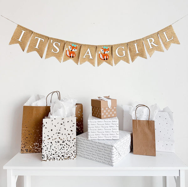 A burlap banner made from 4.5 by 6.5 inch fishtail shaped flags. Each flag has a pocket across the top for stringing. The banner says IT&#39;S A GIRL in white capital letters. There is a hand stenciled image of a fox with an aqua green bow between the words.  It is strung on a jute string that is 110 inches in length.