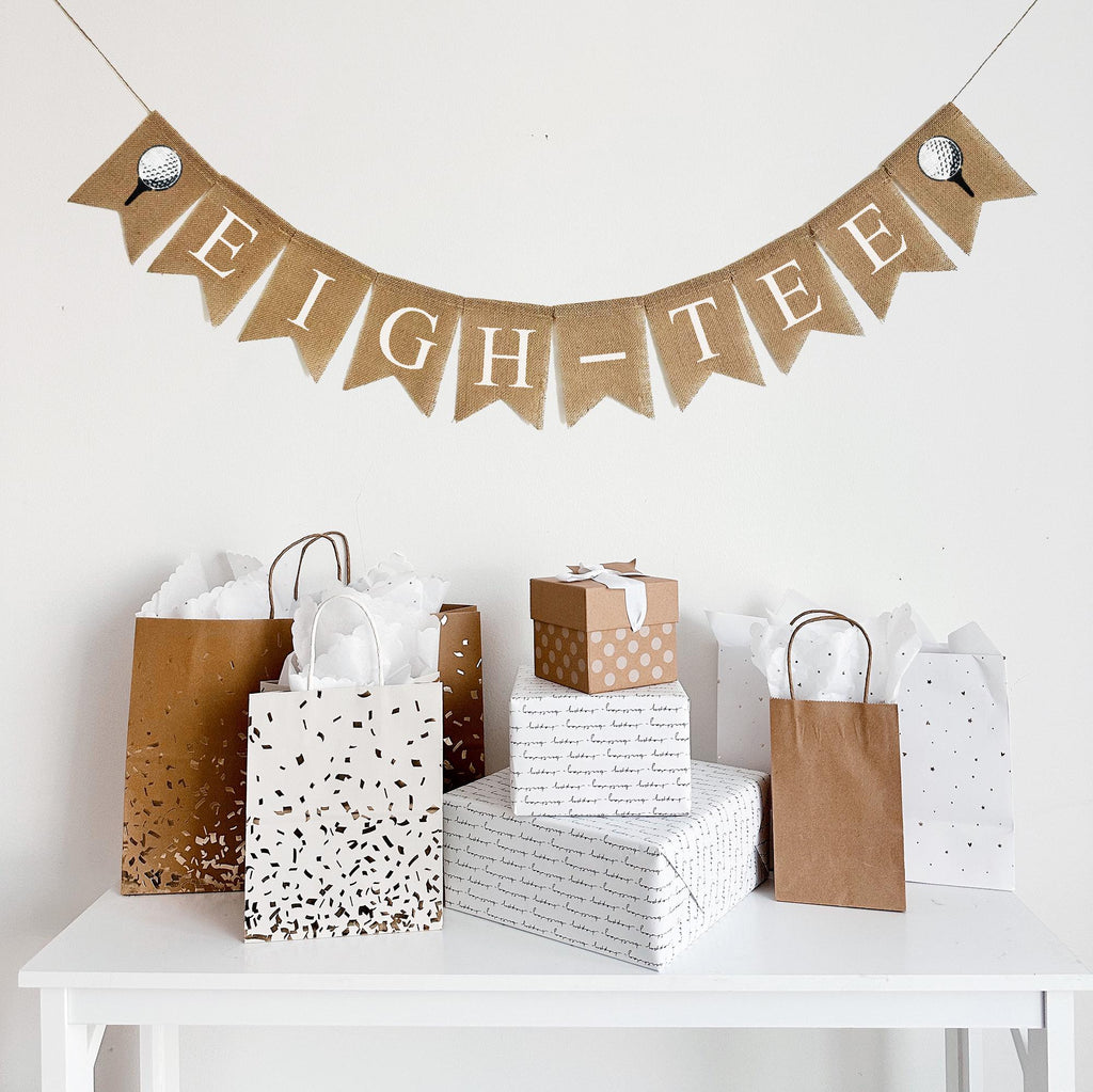 A burlap banner made from 4.5 by 6.5 inch fishtail shaped flags. Each flag has a pocket across the top for stringing. It says EIGH-TEE in white capital letters and has a hand stenciled image of a white golf ball sitting on a black tee on each end. It is strung on a jute string that is 110 inches in length.