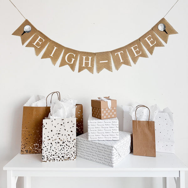 A burlap banner made from 4.5 by 6.5 inch fishtail shaped flags. Each flag has a pocket across the top for stringing. It says EIGH-TEE in white capital letters and has a hand stenciled image of a white golf ball sitting on a black tee on each end. It is strung on a jute string that is 110 inches in length.