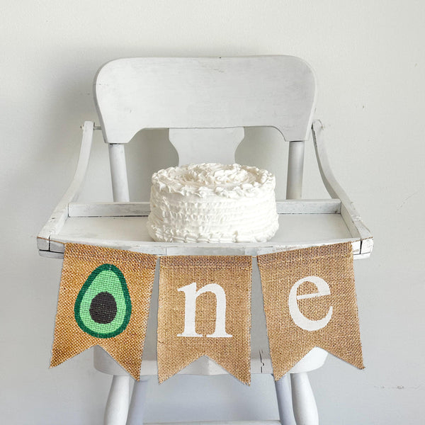 A burlap banner made from three 5 by 7 inch fishtail shaped flags. The first flag has a avocado image on it. The 2nd flag has a white lowercase N, the 3rd flag has a white lowercase letter E. The banner is hung on a high chair in the photo.