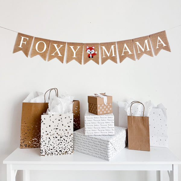 A burlap banner made from 4.5 by 6.5 inch fishtail shaped flags. Each flag has a pocket across the top for stringing. This banner says FOXY MAMA in white capital letters. There is a hand stenciled image of a fox in between the words. It is strung on a jute string that is 110 inches in length.