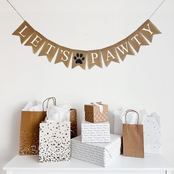 A burlap banner made from 4.5 by 6.5 inch fishtail shaped flags. Each flag has a pocket across the top for stringing. This banner says LET&#39;S PAWTY in white capital letters. There is a hand stenciled image of a black paw print in between the two words. It is strung on a jute string that is 110 inches in length.