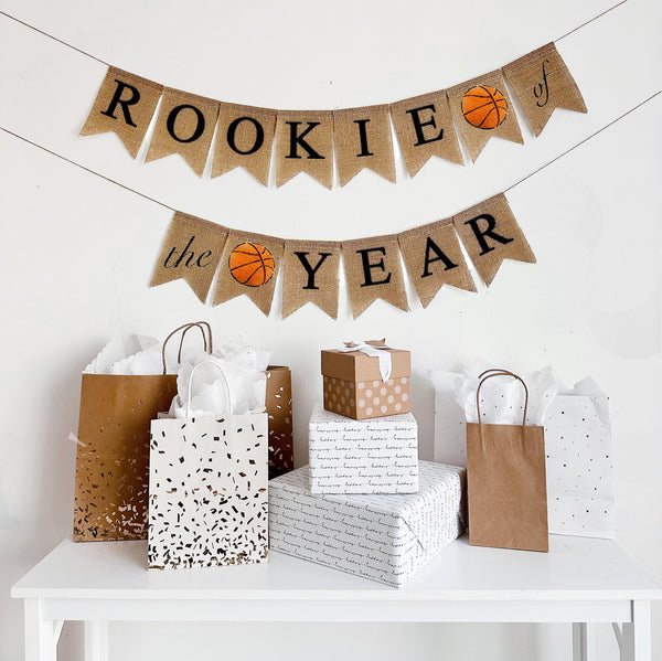 A two tiered burlap banner. The first tier reads ROOKIE OF with a basketball flag between the words and the second tier reads THE YEAR with a basketball between the words.