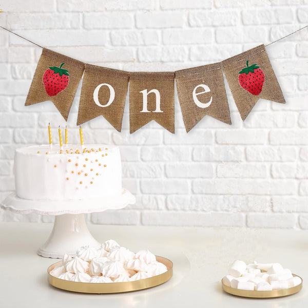 This first birthday banner has 5 burlap flags. It starts with a hand stenciled red strawberry with a green leaf and stem following by three flags spelling out the word ONE in white lowercase letters followed by another hand stenciled strawberry.