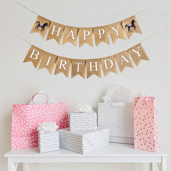 A burlap banner made from 4.5 by 6.5 inch fishtail shaped flags. Each flag has a pocket across the top for stringing. The top line of this banner says HAPPY in white capital letters on the top line with a hand stenciled image of a brown horse with a pink saddle on each end. The bottom line says BIRTHDAY in white capital letters. It is strung on two 110 inch jute strings.