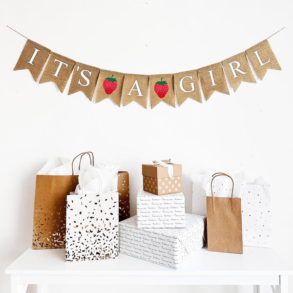 This baby shower banner hangs on one line. The banner reads It&#39;s A Girl in white uppercase letters with a red hand stenciled strawberry in between each word.