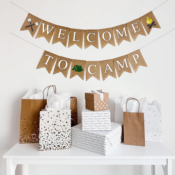 A burlap banner made from 4.5 by 6.5 inch fishtail shaped flags. Each flag has a pocket across the top for stringing. The top line of this banner says WELCOME on the top line in white capital letters. There is a hand stenciled image of crossed sticks with marshmallows on one end and a campfire on the other end. The second line says TO CAMP in white capital letters and has a hand stenciled image of a green tent in between the words. It is strung on two jute strings that are 110 inches in length.