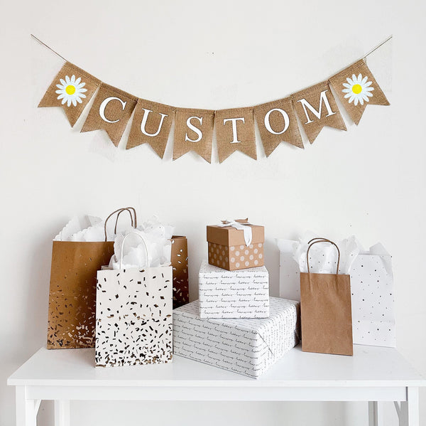 A burlap banner made from 4.5 by 6.5 inch fishtail shaped flags. Each flag has a pocket across the top for stringing. This is a custom banner in white capital letters. Add the name of your choosing to personalize this banner. There is a hand stenciled image of a white daisy with a yellow center on each end of the name. It is strung on a jute string that is 110 inches in length.
