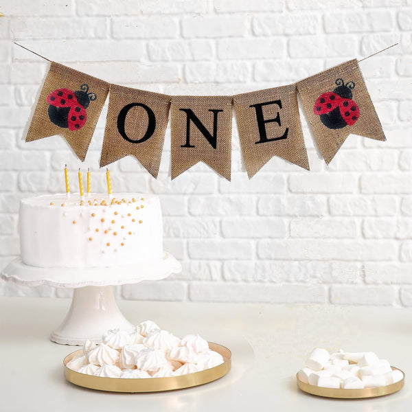 A burlap banner that says ONE in black letters with red and black ladybug images on either side of the word.