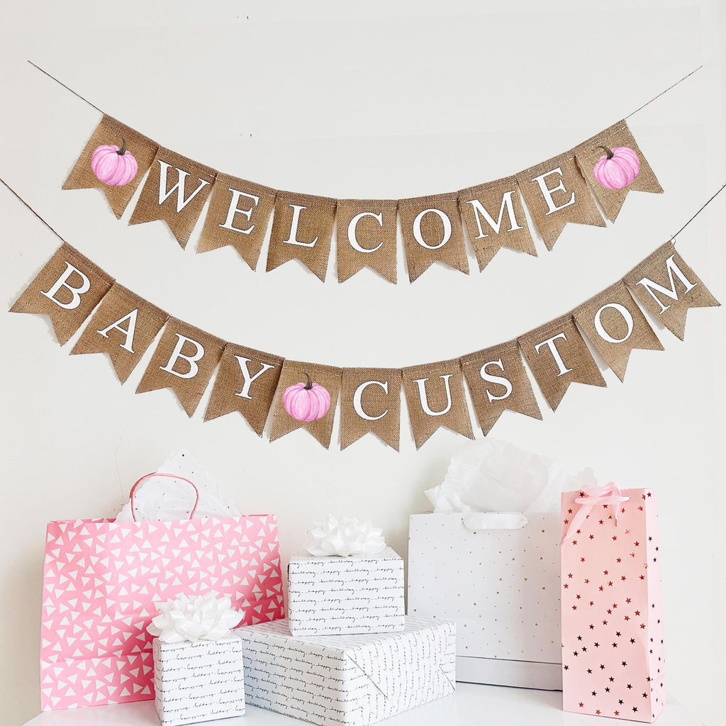A burlap banner made from 4.5 by 6.5 inch fishtail flags. The top line says WELCOME in white capital letters and has a pink pumpkin on each end. The bottom string says BABY with your choice of custom name and has a pink pumpkin in the middle.