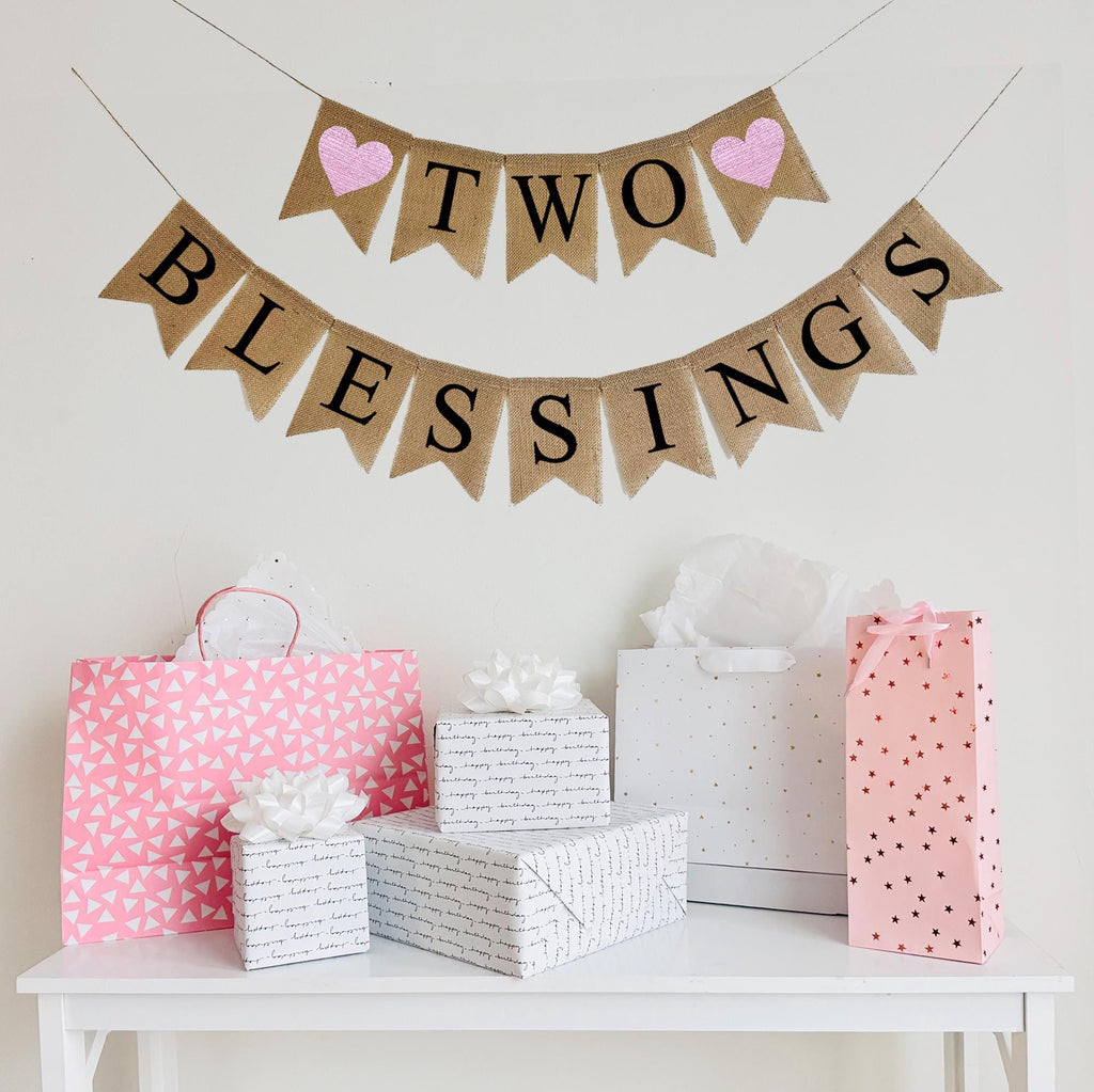A burlap banner made from 4.5 by 6.5 inch fishtail shaped flags. Each flag has a pocket across the top for stringing. The top line says TWO in black capital letters and has a pink heart on each end. The bottom line says BLESSINGS in black capital letters. It is strung on two 110 inch jute strings.