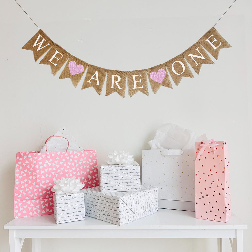 A burlap banner made from 4.5 by 6.5 inch fishtail shaped flags. Each flag has a pocket across the top for stringing. This banner says WE ARE ONE in white capital letters with an image of a pink heart between each of the words. It is strung on a jute string that is 110 inches in length.