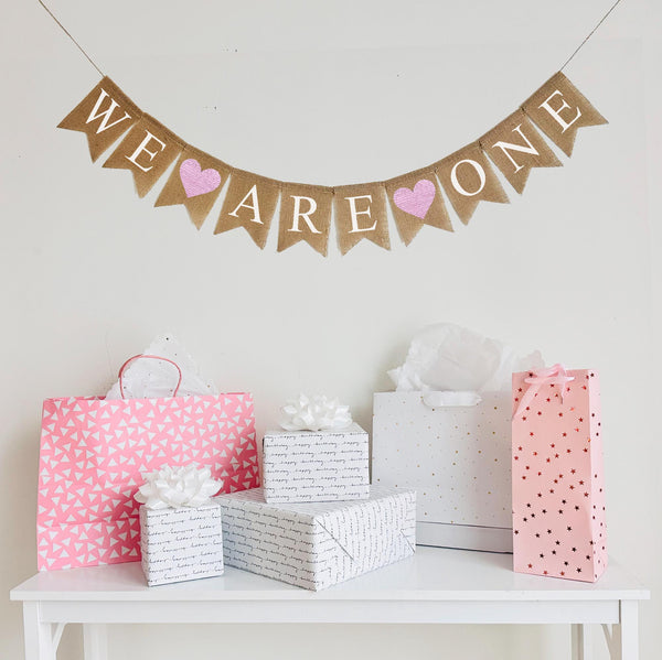 A burlap banner made from 4.5 by 6.5 inch fishtail shaped flags. Each flag has a pocket across the top for stringing. This banner says WE ARE ONE in white capital letters with an image of a pink heart between each of the words. It is strung on a jute string that is 110 inches in length.