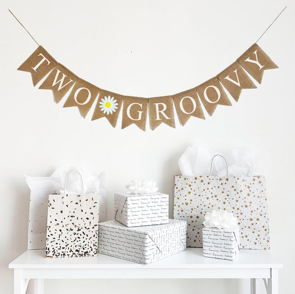 A burlap banner made from 4.5 by 6.5 inch fishtail shaped flags. Each flag has a pocket across the top for stringing. This banner says TWO GROOVY in white capital letters. There is a hand stenciled image of a white daisy with a yellow center in between the two words. It is strung on a jute string that is 110 inches in length.