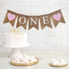 This is a burlap banner made of 5 fishtail shaped flags. The word ONE is spelled out in white capital letters on the middle three flags. There is an image of a pink glitter heart on each end of the banner. The flags measure 4.5 by 6.5 inches each.