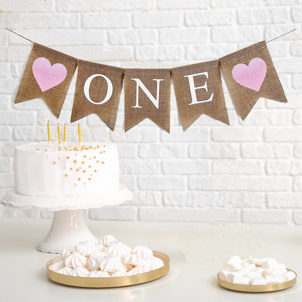 This is a burlap banner made of 5 fishtail shaped flags. The word ONE is spelled out in white capital letters on the middle three flags. There is an image of a pink glitter heart on each end of the banner. The flags measure 4.5 by 6.5 inches each.