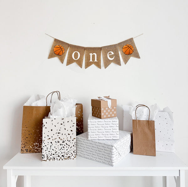A burlap banner that says &quot;one&quot; in white lowercase lettering with orange basketball images on either side of the words.