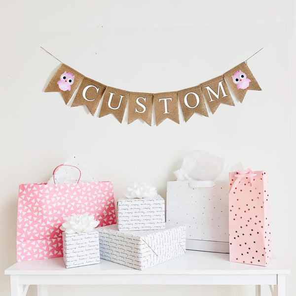 A burlap banner made from 4.5 by 6.5 inch fishtail shaped flags. Each flag has a pocket across the top for stringing. This is a custom banner in white capital letters. Add the name of your choosing to personalize this banner. There is a hand stenciled image of a pink owl on each end of the name. It is strung on a jute string that is 110 inches in length.