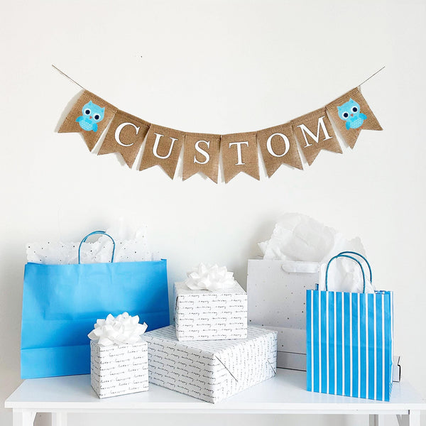 A burlap banner made from 4.5 by 6.5 inch fishtail shaped flags. Each flag has a pocket across the top for stringing. This is a custom banner in white capital letters. Add the name of your choosing to personalize this banner. There is a hand stenciled image of a blue owl on each end of the name. It is strung on a jute string that is 110 inches in length.