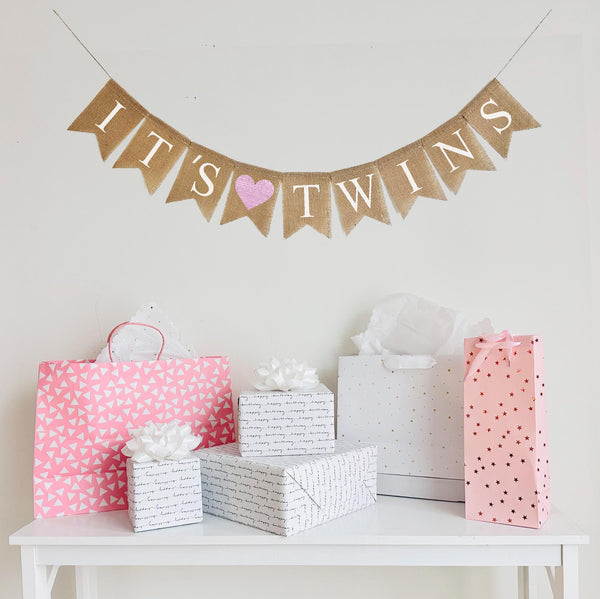 A burlap banner made from 4.5 by 6.5 inch fishtail shaped flags. Each flag has a pocket across the top for stringing. This banner says IT&#39;S TWINS in white capital letters. There is an image of a light pink heart in between the words. It is strung on a 110 inch jute string.