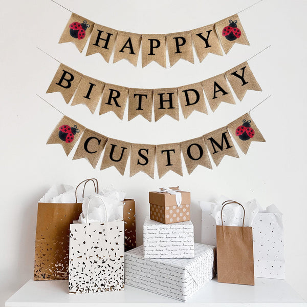 A happy birthday burlap banner strung on three strings. The happy is at the top, birthday second, and custom on the third row. Hand painted red & black ladybugs are at the beginning and end of the word happy and custom.