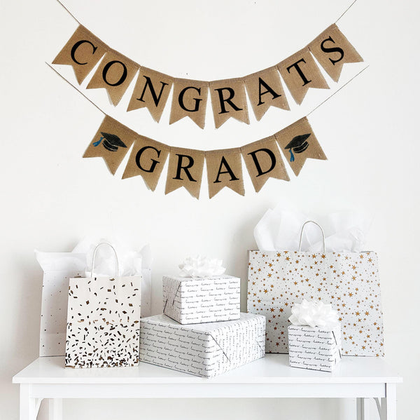 A burlap banner made from 4.5 by 6.5 inch fishtail shaped flags. Each flag has a pocket across the top for stringing. The top line says CONGRATS in black capital letters. The bottom line says GRAD in black capital letters. There is a hand stenciled image of a black graduation cap with a blue tassel on each end of the word GRAD. Each line is strung on a jute string that is 110 inches in length.