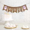 A burlap banner that says &quot;one&quot; in white lowercase lettering with pink owl images on either side of the words.