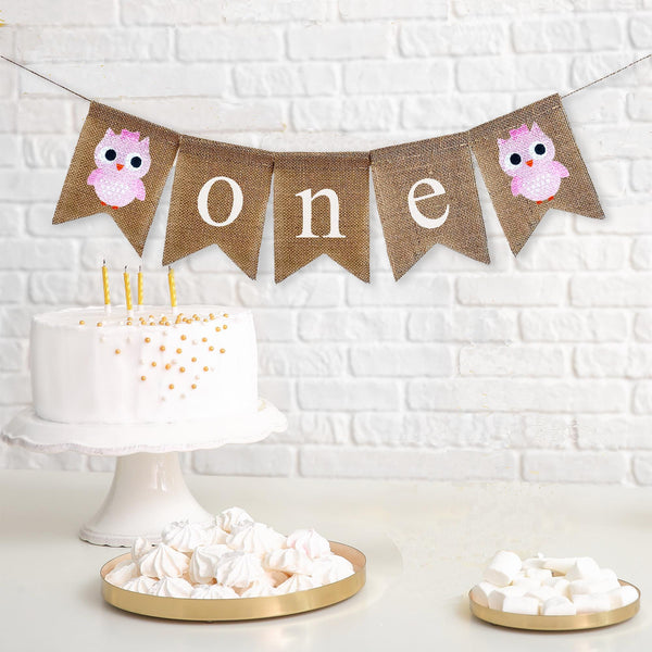 A burlap banner that says &quot;one&quot; in white lowercase lettering with pink owl images on either side of the words.