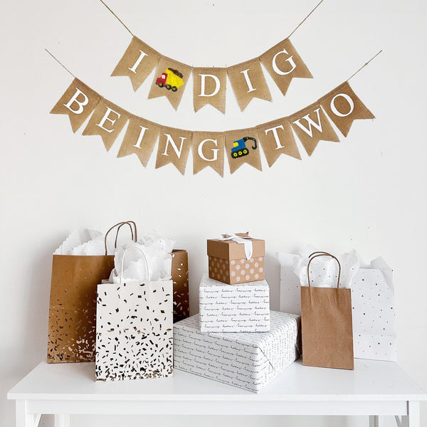 This two tiered banner spells out the words I Dig Being Two. Between the words I and Dig there is a red and yellow dump truck image. The second line reads Being Two with a blue digger truck in between the words Being and Two.